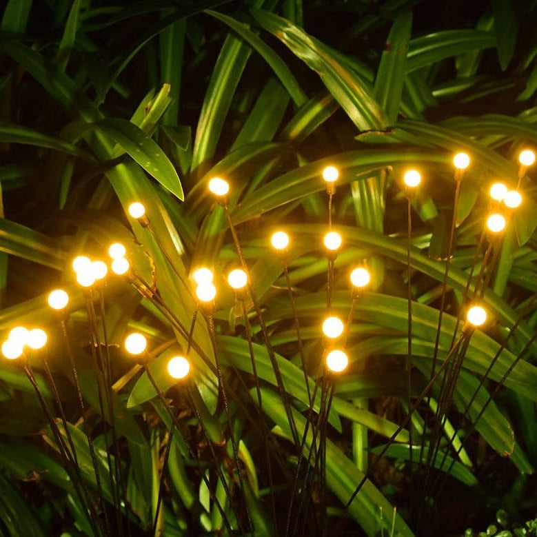 Soft, warm LED stake lights in a garden setting, designed to mimic fireflies glowing among lush green leaves. Perfect for outdoor ambiance, these flexible solar-powered lights blend seamlessly into natural surroundings, providing gentle illumination for pathways, flower beds, and landscaped areas, enhancing the night-time beauty of any garden or patio space.